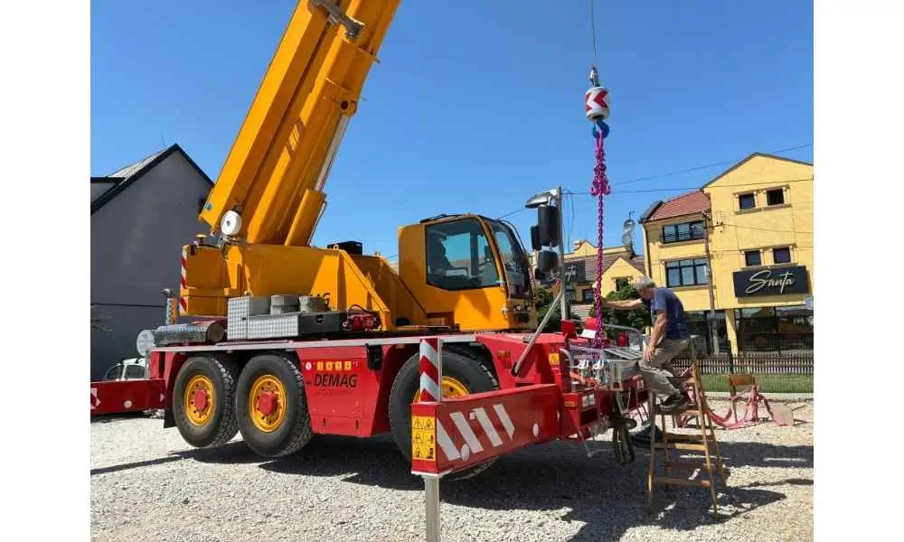 Operater na ljestvici priprema kuku, lance i opremu za dizanje na autodizalici Demag AC 40 City sa izvučenim stabilizatorima na šljunčanom gradilištu u urbanom okruženju — usluge mobilnog krana 40 t Metal Pro